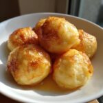 Close-up of pan-fried potato dumplings with a golden-brown crust, sitting in a small pool of sauce in a white bowl.