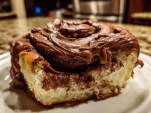 Close-up of a single, soft chocolate cinnamon roll heavily topped with thick, melted chocolate frosting.
