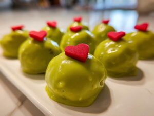 Several bright green Grinch Oreo balls topped with small red candy hearts arranged on a white platter.