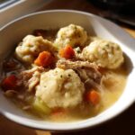 A close-up of a comforting bowl of chicken and dumpling soup with shredded chicken, carrots, and herbs.