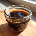 A close-up of rich, dark chocolate syrup filling a small glass jar, sitting on a wooden surface.