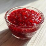 Close-up of vibrant, textured raspberry jam packed with seeds in a small clear glass bowl.