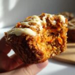 Close-up of a hand holding a bitten carrot cake muffin, showing the moist texture and cream cheese frosting.