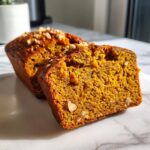 Close-up of a slice of moist pumpkin bread, showing its rich orange color and visible chopped walnuts, sitting next to the rest of the loaf.
