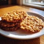 A stack of crisp, golden brown oatmeal lace cookies resting on a white plate, illuminated by bright window light.