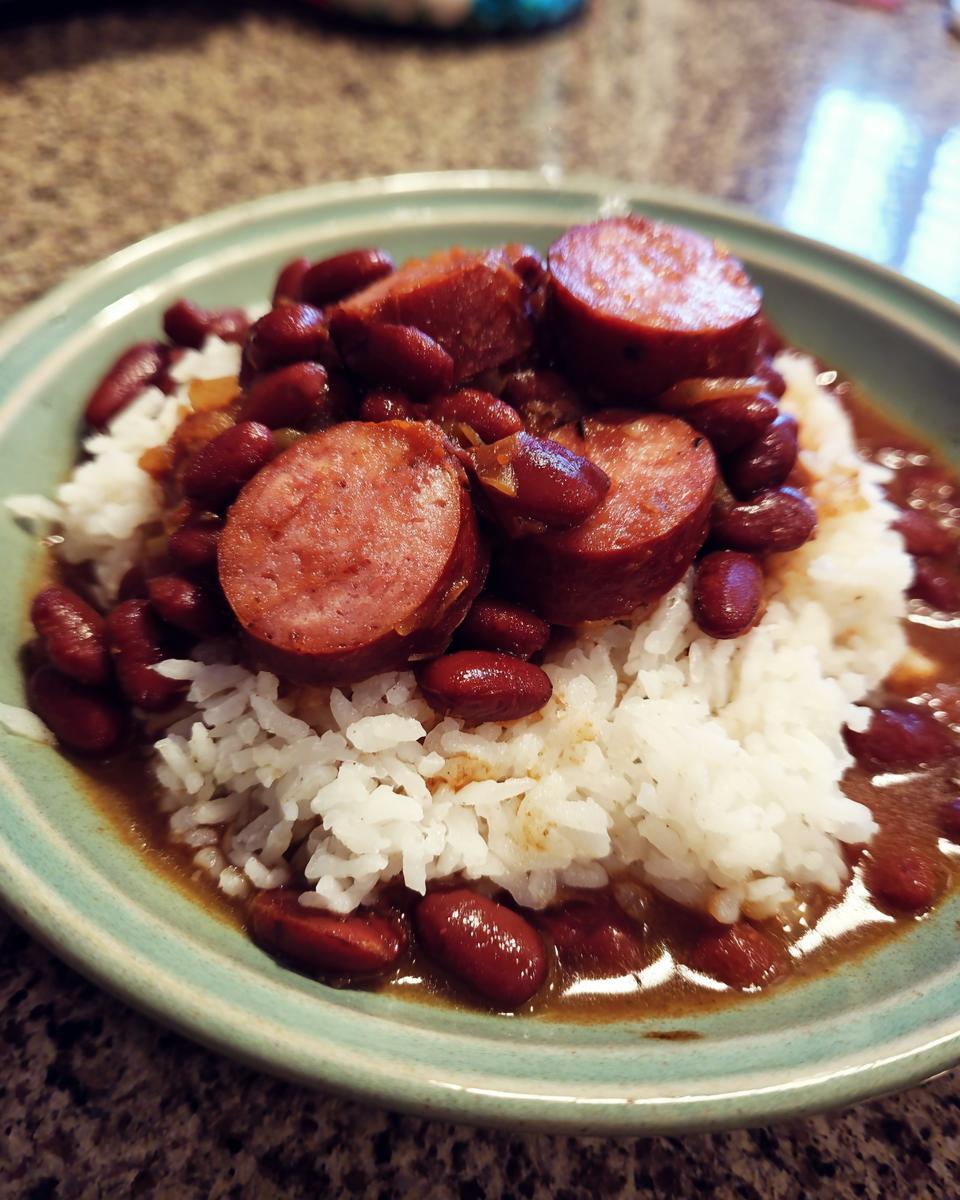 A close-up of a bowl of white rice topped with red kidney beans and thick slices of smoked sausage, representing delicious beans and rice.