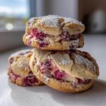 Close-up of stacked raspberry cheesecake cookies filled with pink cream and raspberries, dusted with powdered sugar.