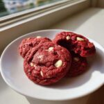 Four vibrant red velvet cake mix cookies with white chocolate chips piled on a white plate near a window.