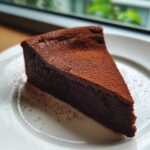A close-up of a dense, dark slice of flourless chocolate torte dusted generously with cocoa powder on a white plate.