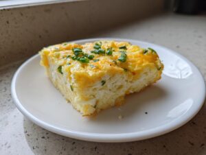 A square slice of fluffy sheet pan eggs topped with melted cheddar cheese and fresh green chives on a white plate.