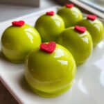 Close-up of several shiny, bright green Grinch Oreo Balls topped with small red, sparkly hearts on a white plate.