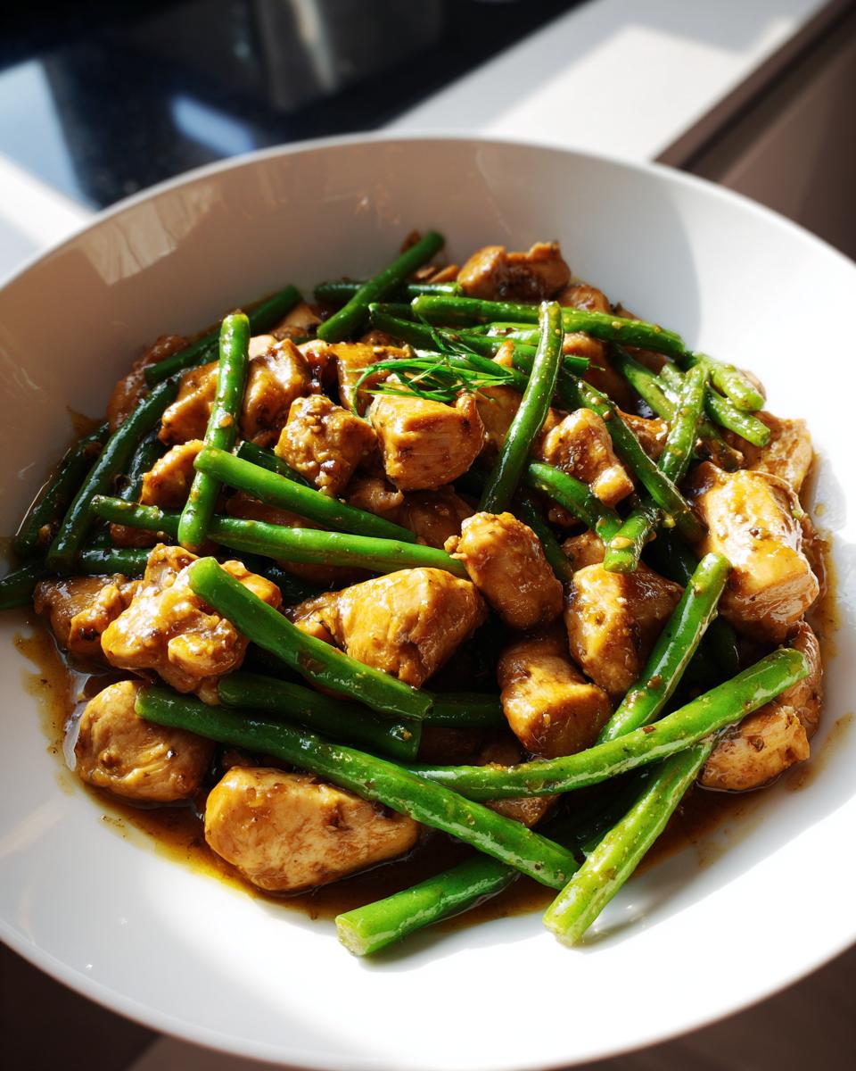 Close-up of bite-sized chicken pieces mixed with bright green beans coated in a savory brown sauce, served in a white bowl.