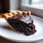 Close-up of a rich, dark slice of fudge pie on a white plate, showing the gooey filling and flaky crust.