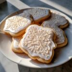 Close-up of sugar cookies topped with smooth, white cookie icing decorated with delicate snowflake patterns.