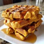 A tall stack of golden orange pumpkin waffles generously drizzled with maple syrup on a white plate.