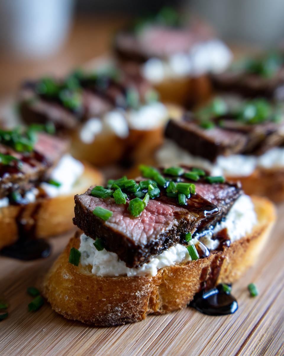 A close-up of one perfect steak crostini topped with creamy cheese, medium-rare steak, balsamic glaze, and fresh chives.