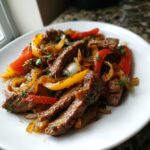 Close-up of delicious steak fajitas featuring sliced seasoned beef, red and yellow bell peppers, and onions on a white plate.