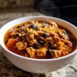 Close-up of a steaming white bowl filled with rich, meaty pumpkin chili topped with melted cheese and black beans.