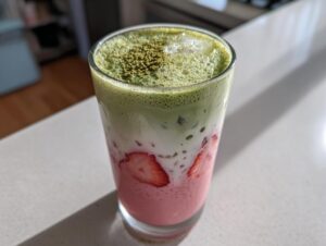 Close-up of a layered strawberry matcha drink featuring pink strawberry puree, white milk, and green matcha foam with ice.