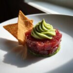 A beautifully plated serving of tuna tartare, cubed and stacked, topped with a swirl of green avocado cream and served with crispy chips.