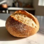 A round, golden-brown loaf of artisan bread, scored on top to reveal a fluffy interior, perfect for making bread bowls.