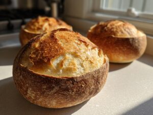 Close-up of three golden-brown, sturdy bread bowls with scored tops sitting on a countertop in sunlight.