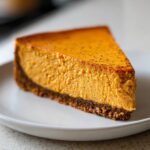 A close-up shot of a thick slice of bright orange sweet potato cheesecake with a dark graham cracker crust on a white plate.
