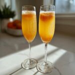 Two flutes filled with a fizzy, orange-colored bellini recipe cocktail sitting on a sunlit marble counter.