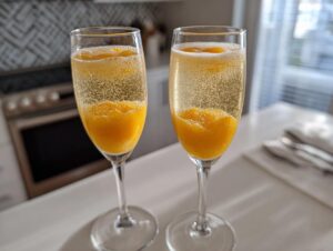 Two champagne flutes filled with a bubbly, sparkling bellini recipe cocktail featuring bright orange peach puree at the bottom.