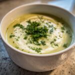 Close-up of a bowl of velvety celery soup, swirled with cream and topped with fresh chopped parsley.