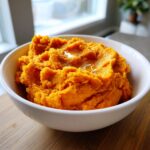 A close-up of fluffy, bright orange mashed sweet potatoes topped with melted butter in a white bowl.