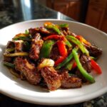 A close-up of a white bowl filled with savory pepper steak, featuring beef strips coated in dark sauce mixed with bright red and green bell peppers.