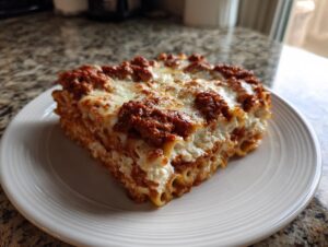 A thick, square slice of baked ziti layered with ricotta, meat sauce, and melted mozzarella cheese on a white plate.