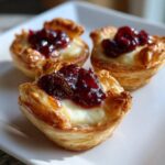 Three golden baked brie bites made with flaky pastry, melted brie cheese, and topped with dark red jam.