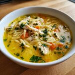 Close-up of a white bowl filled with amazing chicken orzo soup, featuring shredded chicken, carrots, and fresh parsley.