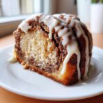 A close-up of a moist slice of cinnamon roll cake recipe topped with thick white vanilla icing dripping down the sides.