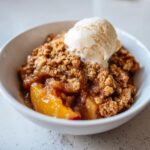Close-up of a warm serving of amazing peach crisp topped with a scoop of vanilla ice cream in a white bowl.