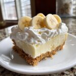 A square slice of banana cream cheesecake with a graham cracker crust, topped with whipped cream and fresh banana slices.