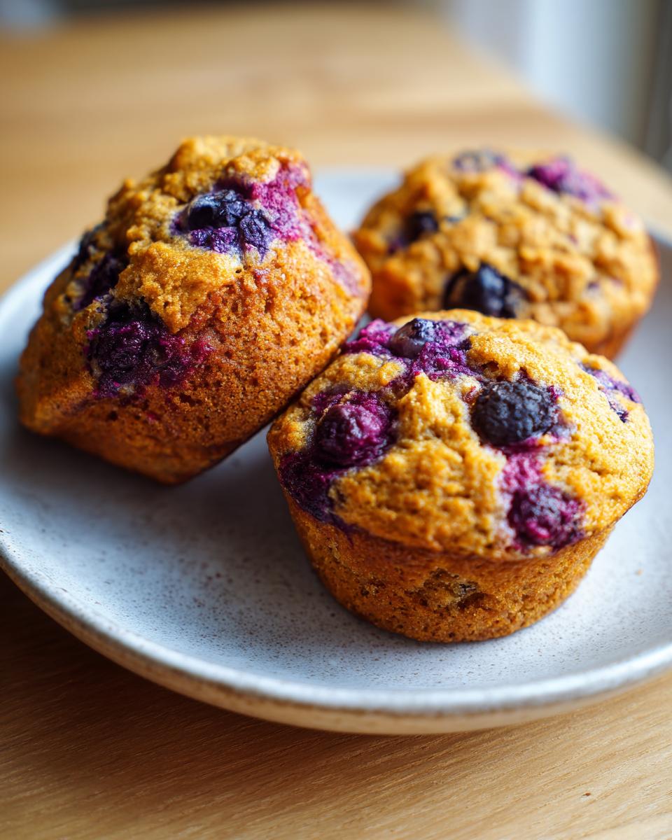 Three golden-brown blueberry cottage cheese muffins with visible purple berries resting on a speckled ceramic plate.