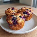 Three golden-brown blueberry cottage cheese muffins with bursting purple berries on a speckled white plate.
