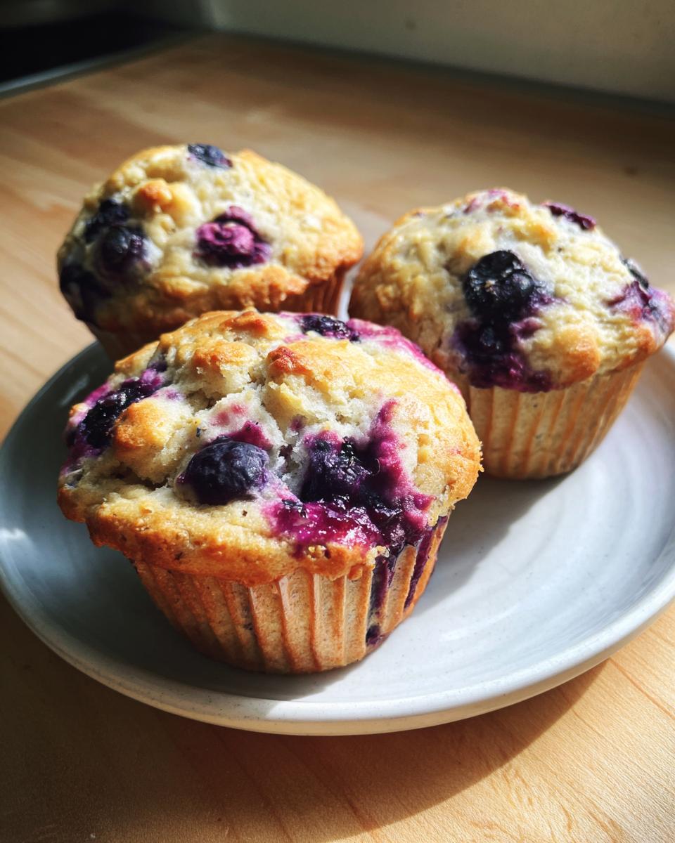Three golden blueberry cottage cheese muffins sitting on a light gray plate on a wooden surface.