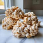 A stack of four homemade avalanche cookies, coated in melted marshmallow and cereal, sitting on a white marble surface.