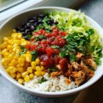 A vibrant, close-up view of a fully loaded chicken burrito bowl featuring rice, shredded chicken, corn, black beans, salsa, and lettuce.