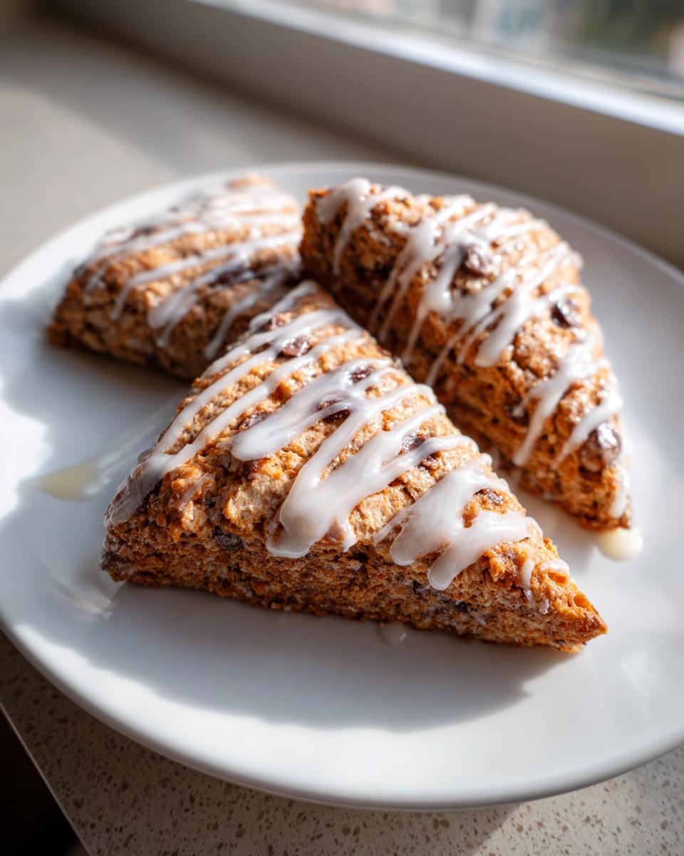 Three triangular chocolate chip scones drizzled with white vanilla glaze, resting on a white plate near a window.