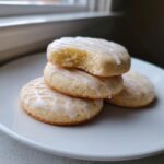 A stack of four soft, glazed london fog cookies on a white plate, with the top cookie having a bite missing.