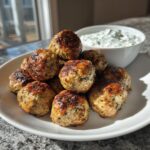 A pile of golden brown greek chicken meatballs served on a white plate next to a bowl of tzatziki sauce.