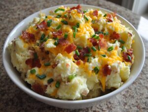 A close-up of a bowl filled with creamy baked potato salad topped with shredded cheddar cheese, bacon bits, and chopped chives.