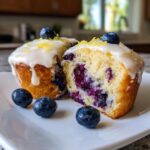 One lemon blueberry cupcake cut in half showing moist interior packed with blueberries, topped with lemon glaze.