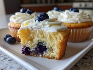 Close-up of a lemon blueberry cupcake cut in half showing moist cake, baked blueberries, and white frosting topped with fresh blueberries.