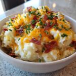 Close-up of a bowl filled with creamy baked potato salad topped with melted cheddar cheese, bacon bits, and fresh chives.